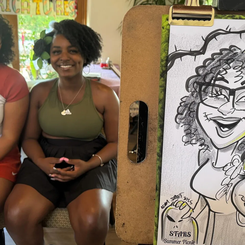 Smiling guest seated beside her Adam Street caricature on the easel at the STARS Summer Picnic